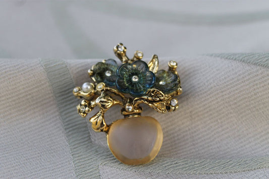 A hyper-realistic editorial photograph capturing the exquisite 'Vaso dell’Anima' brooch from the 1980s, adorned with glistening zircons and luminous pearls, set against a backdrop of rich textures. The brooch is elegantly displayed on a dee