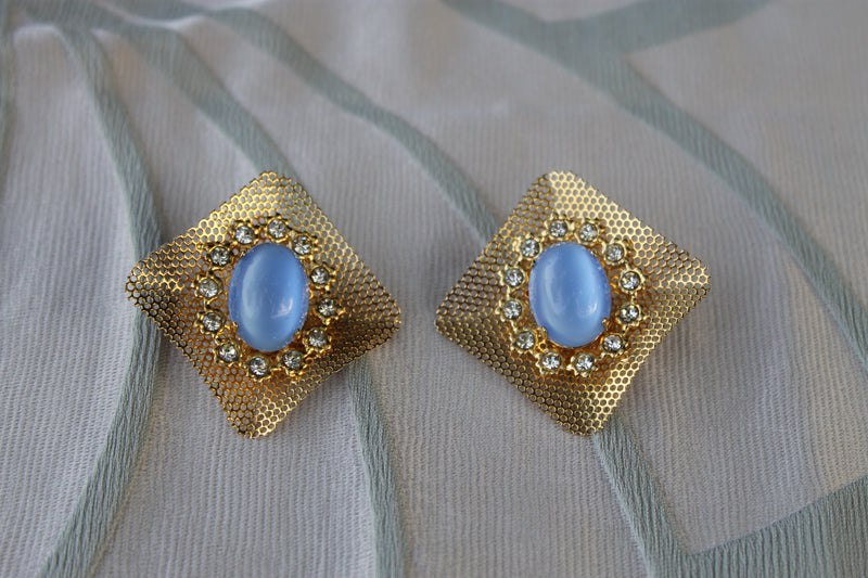 A hyper-realistic editorial photograph featuring the Reticolo di Luce earrings, elegantly displayed against a backdrop of rich textures. The earrings, cast in gold-tone metal and adorned with serene light blue cabochons, catch the soft morn