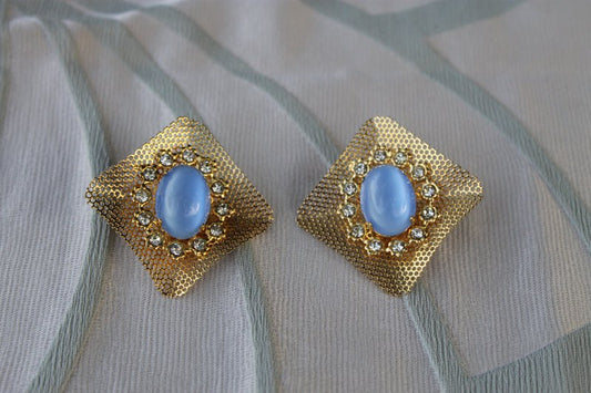 A hyper-realistic editorial photograph featuring the Reticolo di Luce earrings, elegantly displayed against a backdrop of rich textures. The earrings, cast in gold-tone metal and adorned with serene light blue cabochons, catch the soft morn